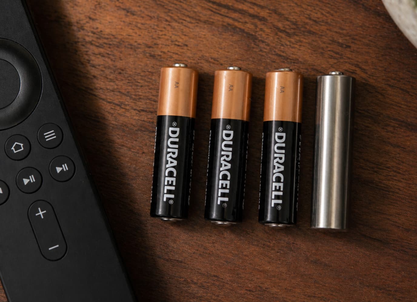 Keepcrypt Cell beside three real AA cells next to a remote on a wood surface, shot identically